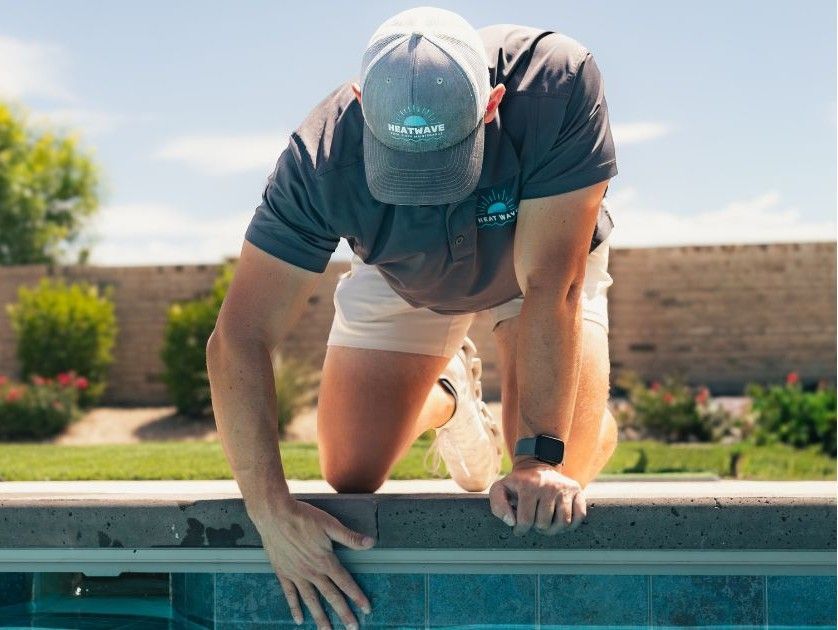 Person kneeling on a pool edge, reaching down to inspect or fix the waterline tile.