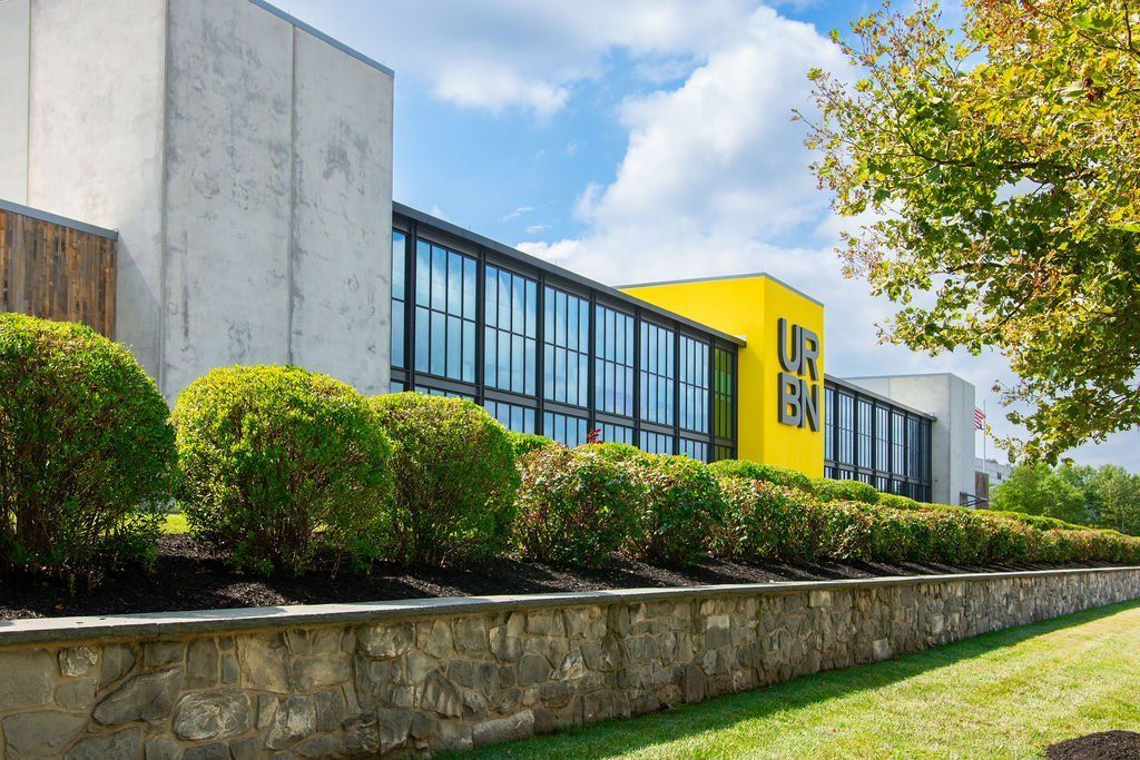 A modern building with large windows and a yellow facade, viewed behind a low stone wall and row of manicured shrubs.