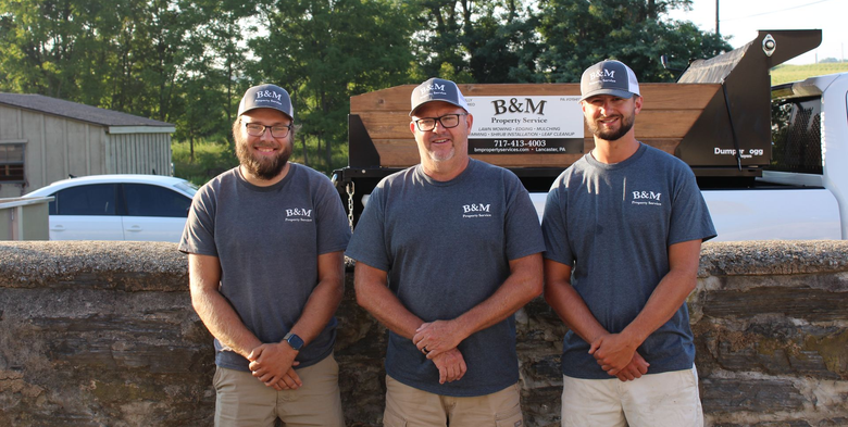 Four people in grey tank tops and khaki shorts stand in a row outdoors in front of green bushes and a tree.