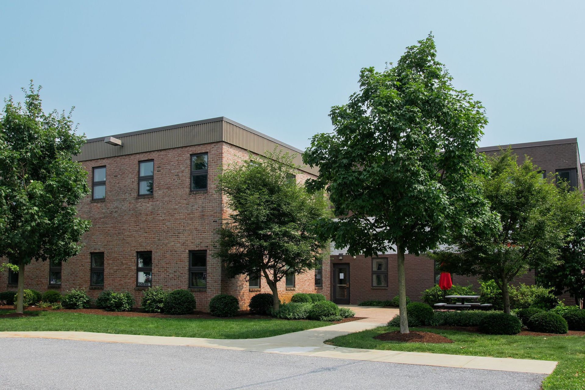 A two-story brick office building framed by lush green trees on a sunny day with a paved walkway in the foreground.