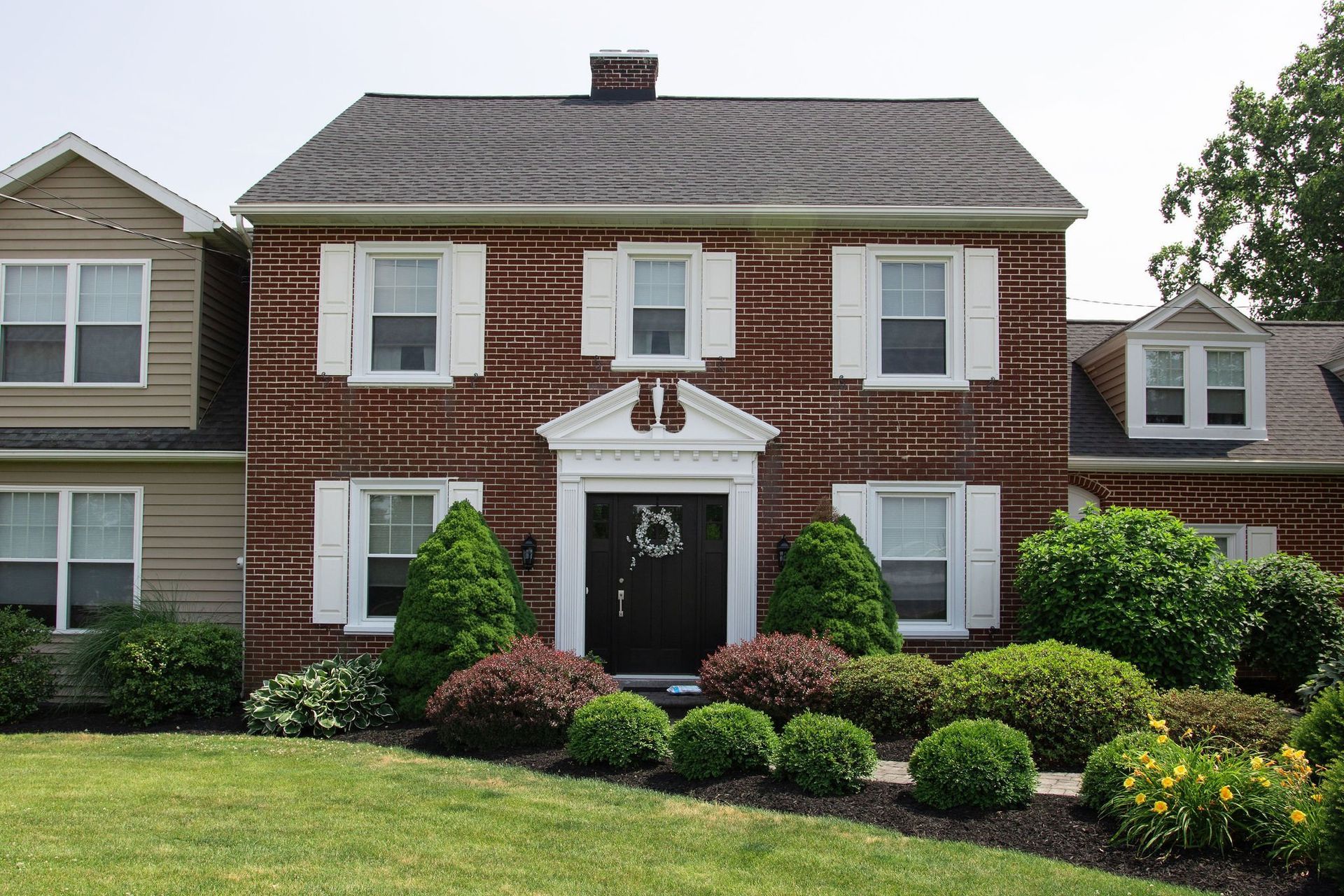 A colonial-style brick house with white shutters, a dark front door, and neatly landscaped shrubs in front.