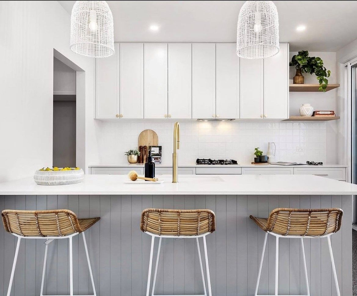 Modern white kitchen with island, rattan bar stools, pendant lights, and open shelving with plants.