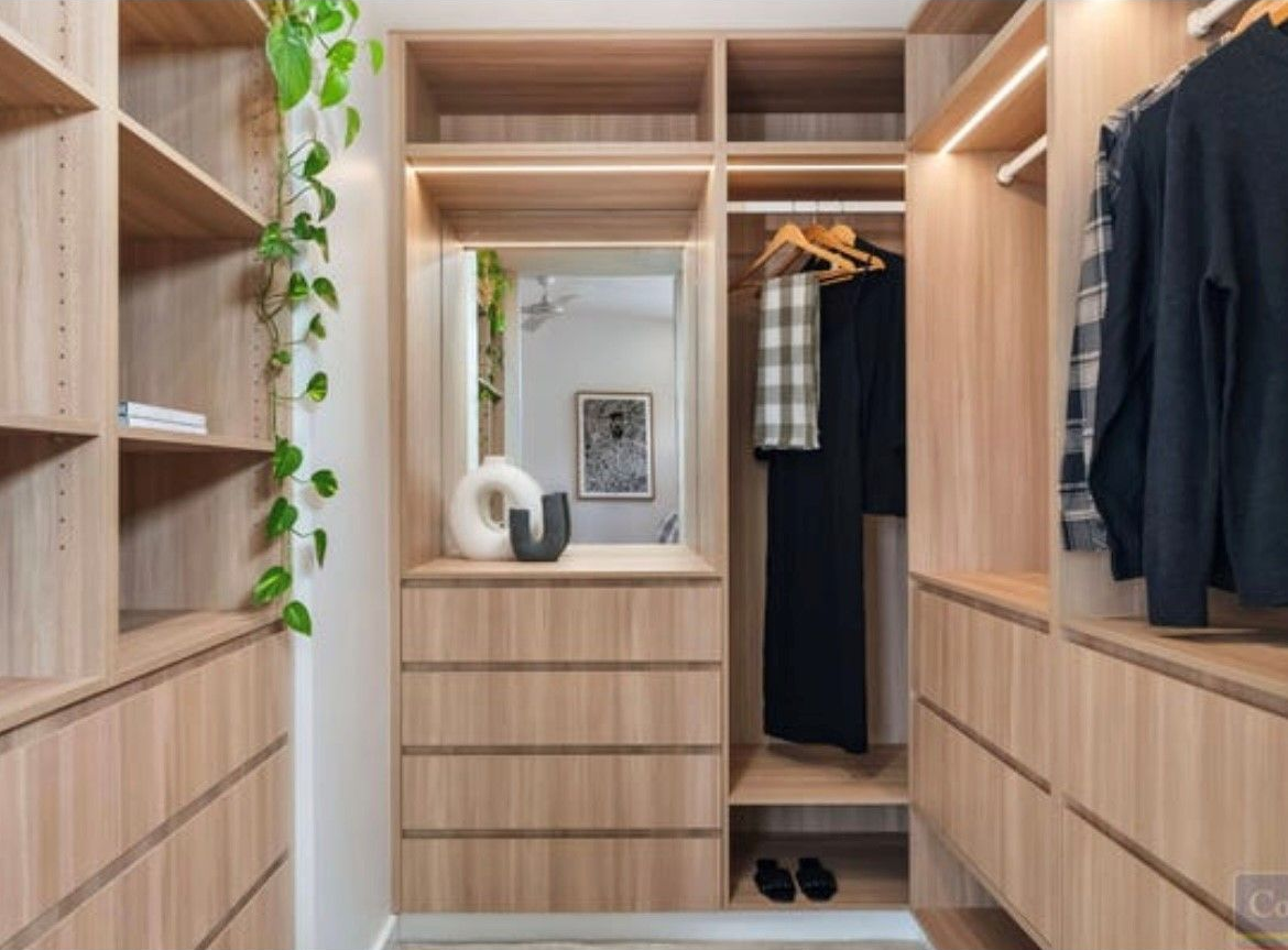Walk-in closet with light wood shelving and drawers, featuring a mirror, hanging clothes, and decorative objects — Inovative Interiors In Cardiff, NSW