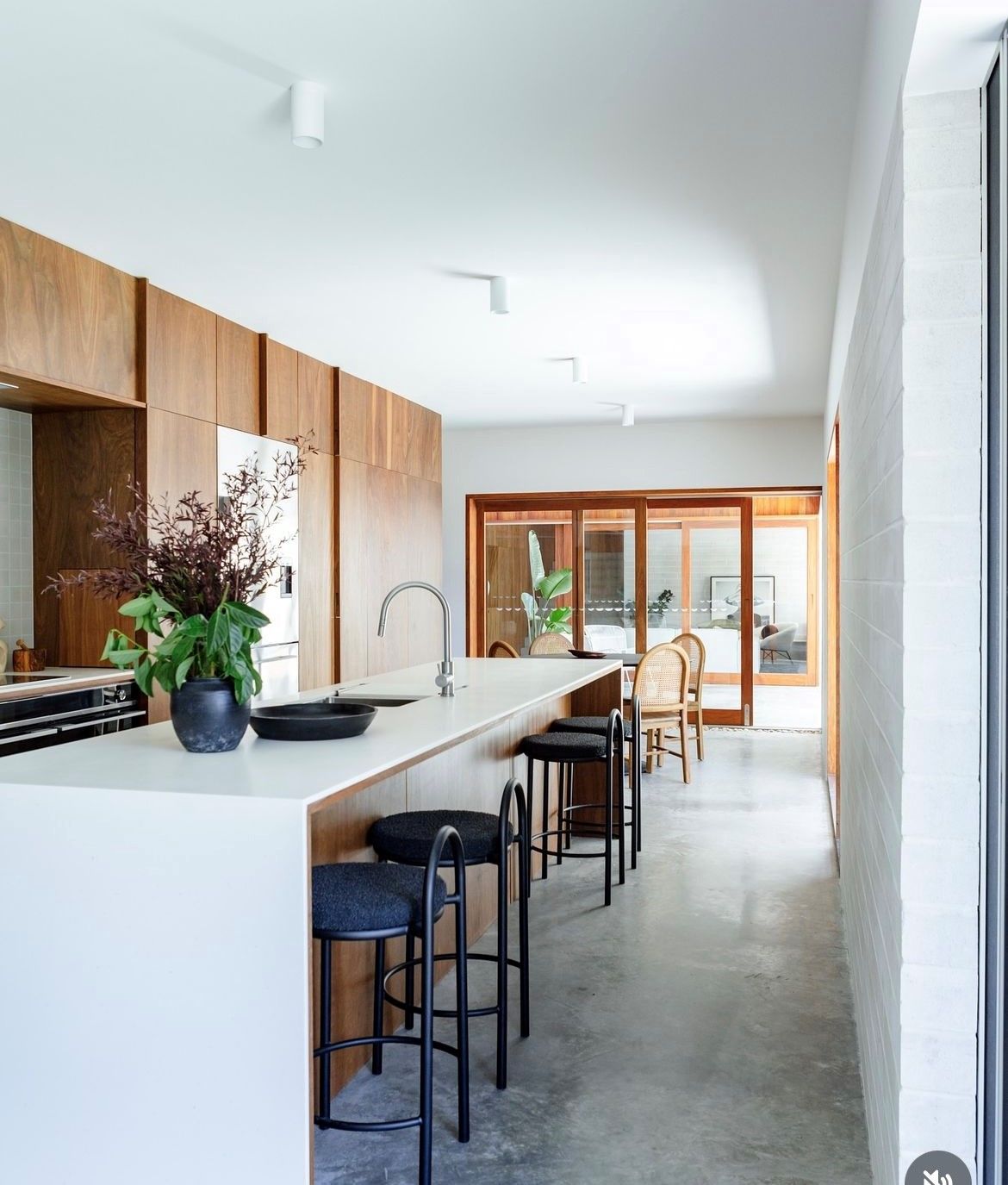 A modern kitchen with a long white island, wooden cabinets, and black bar stools. Natural light streams in through large windows and sliding doors — Inovative Interiors In Cardiff, NSW