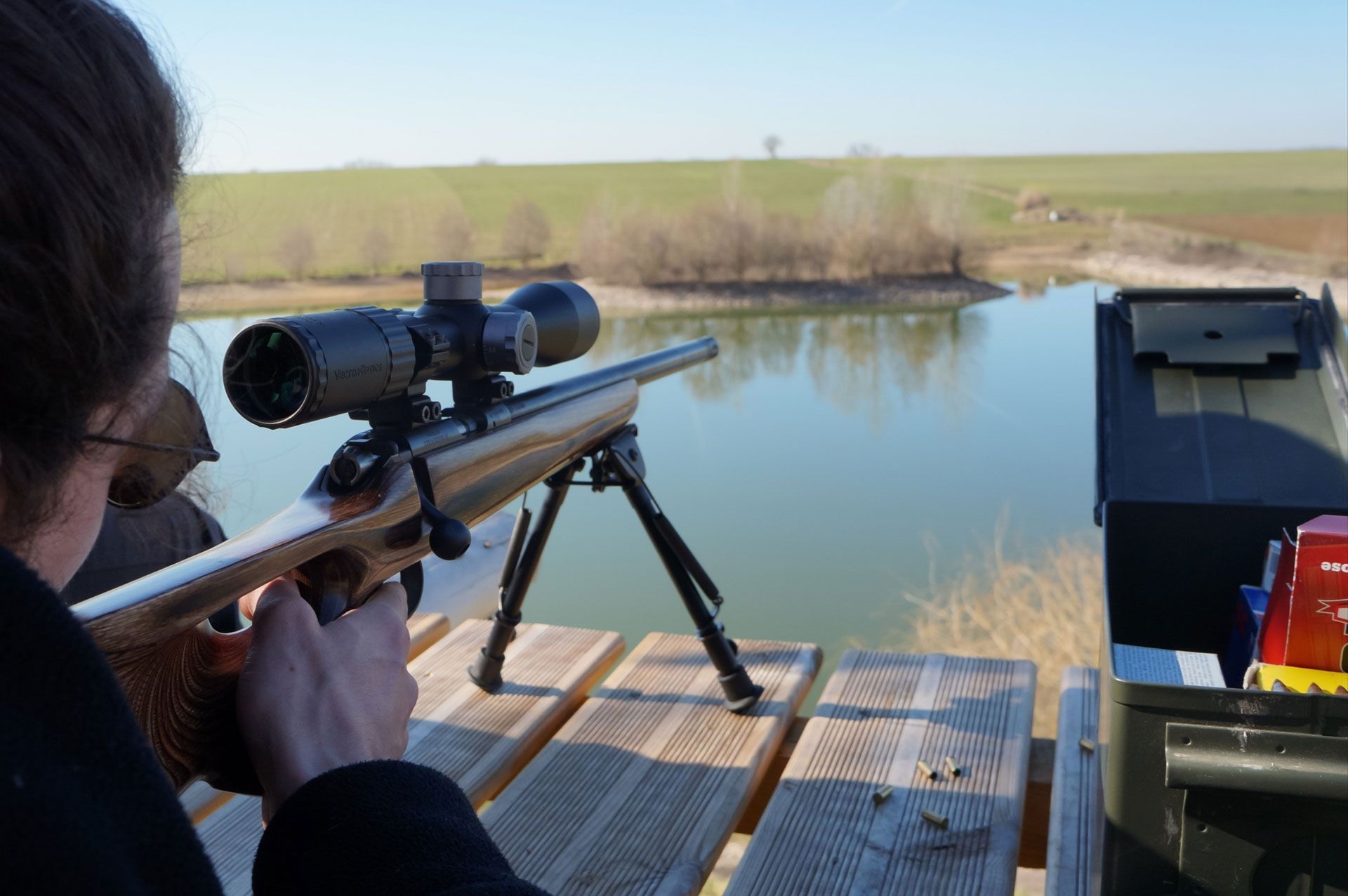 Person aiming a rifle with a scope, mounted on a bipod, at a pond in a sunny outdoor setting.