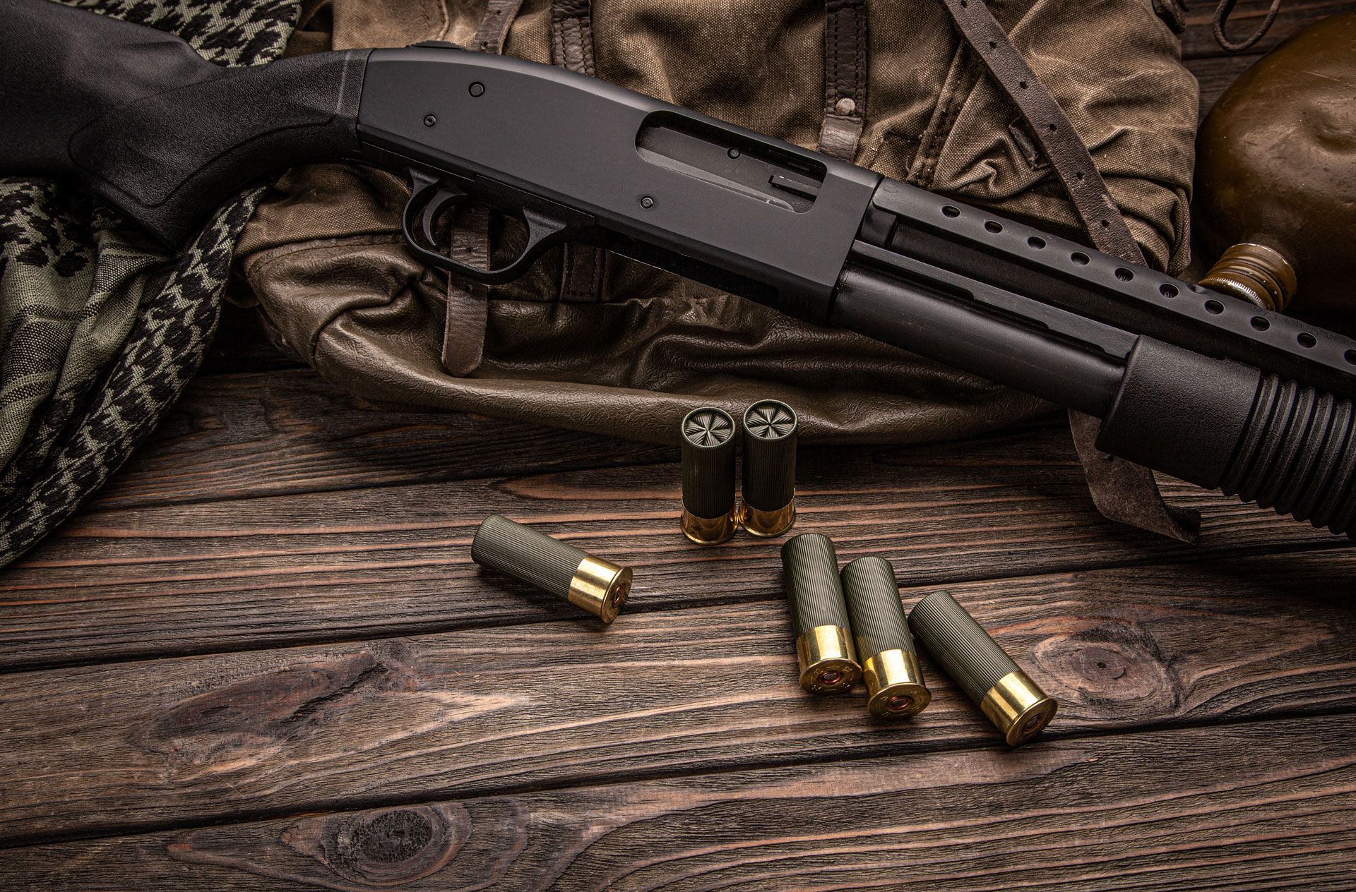 Shotgun and shells on wooden surface, with a backpack and canteen in the background.
