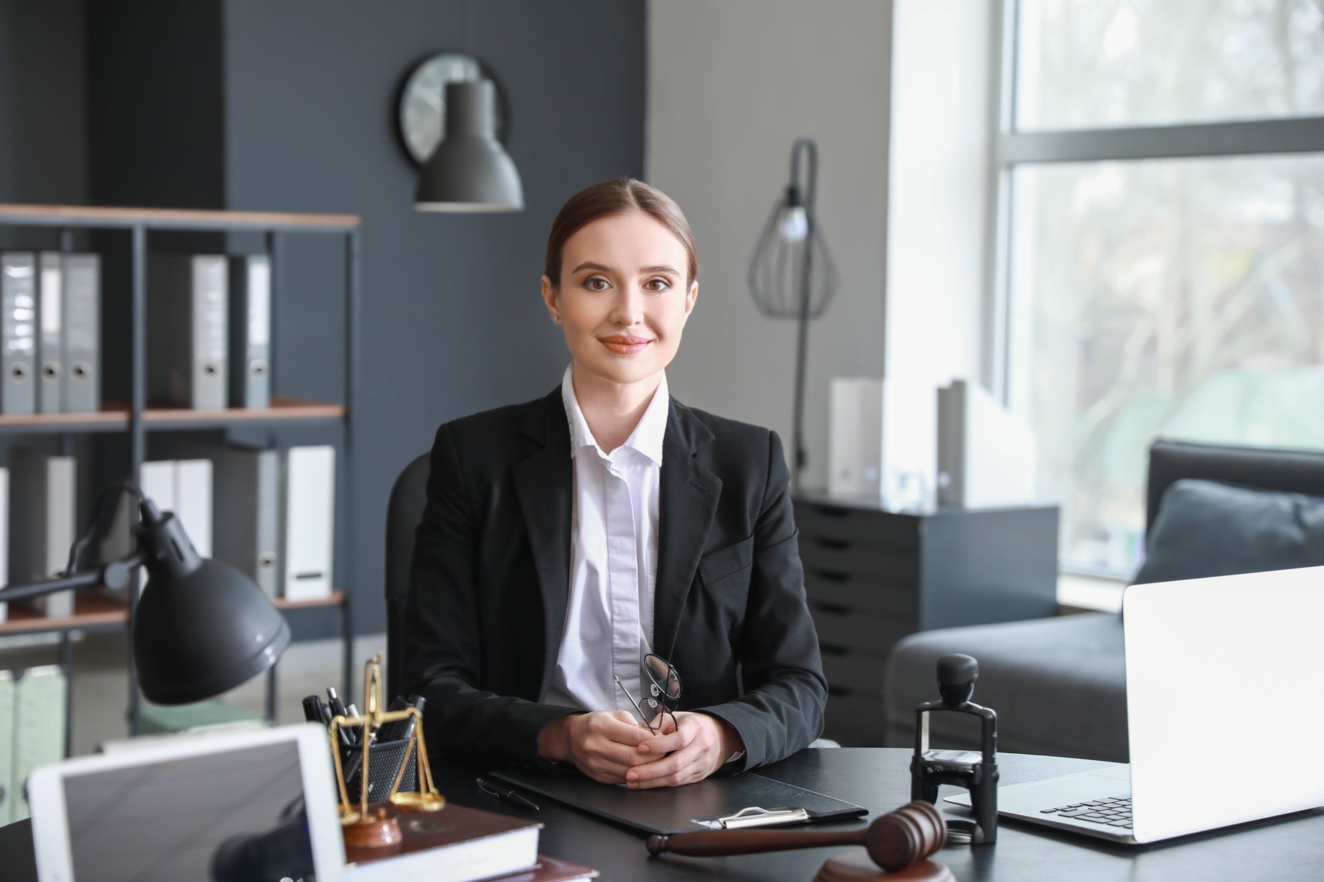 Professional in black blazer seated at an office desk, with laptop, files, and desk lamp in a bright modern workspace.