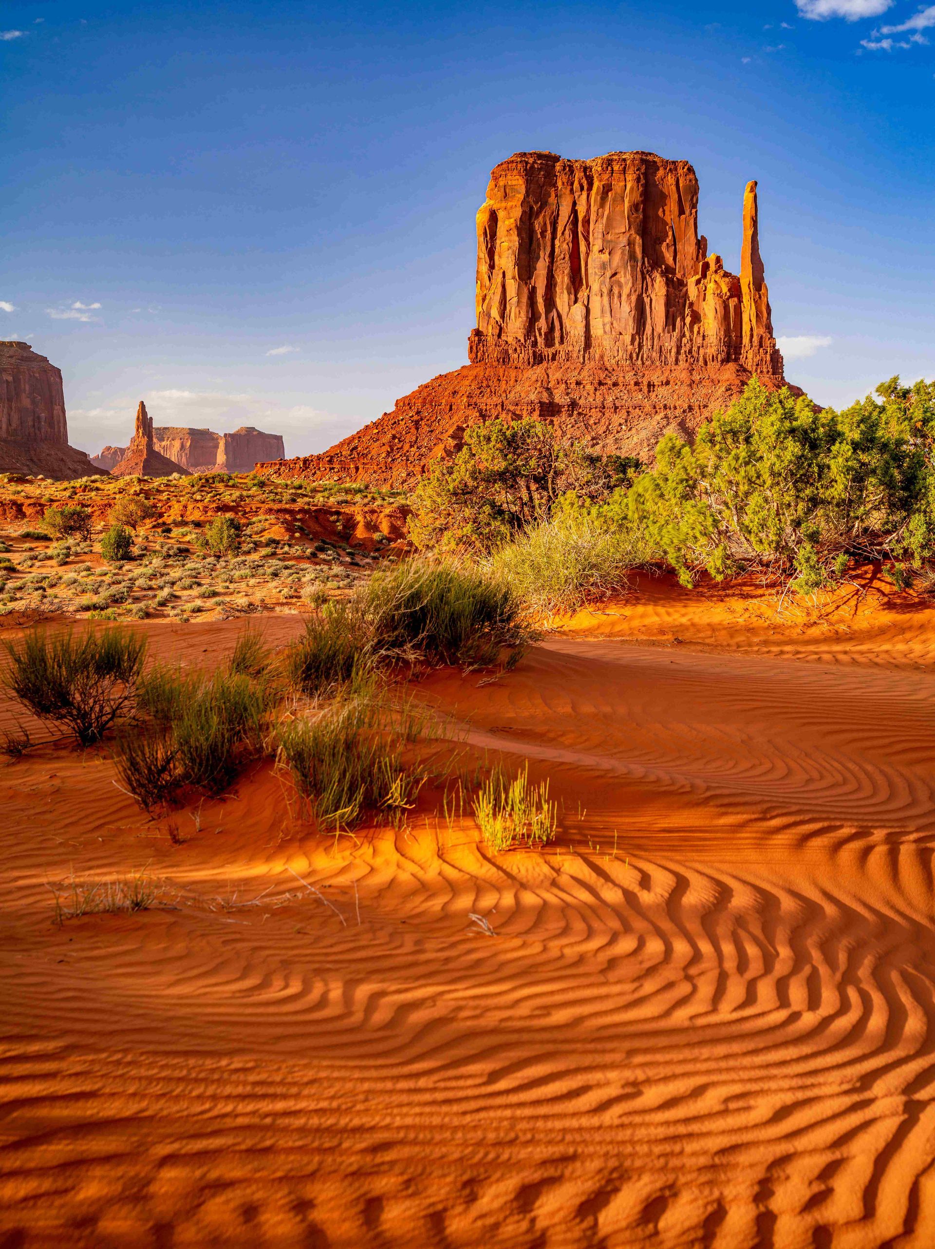 Red rock buttes rising above rippled orange sand in a desert landscape under a blue sky