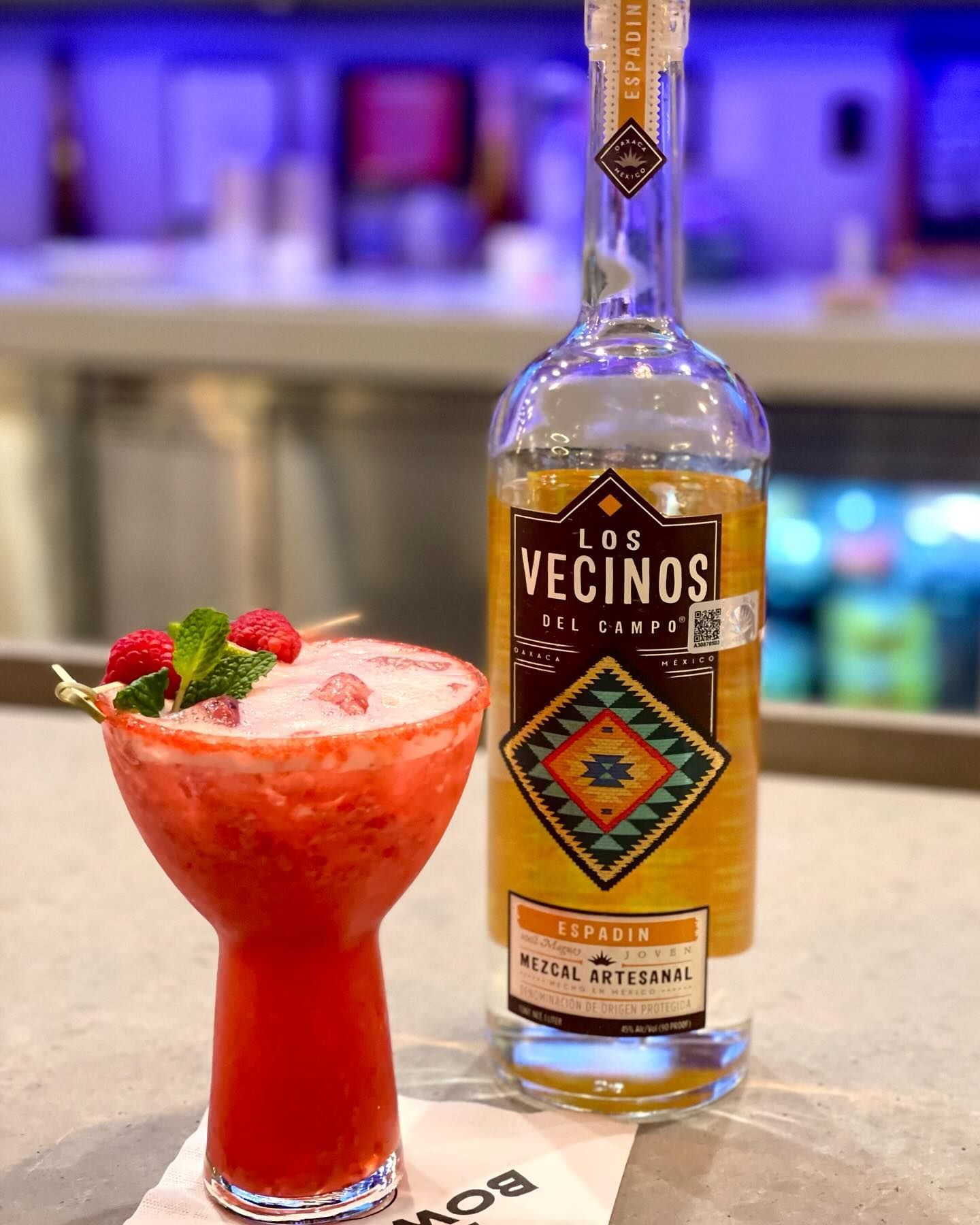 Red cocktail with strawberries beside a Los Vecinos bottle on a bar counter
