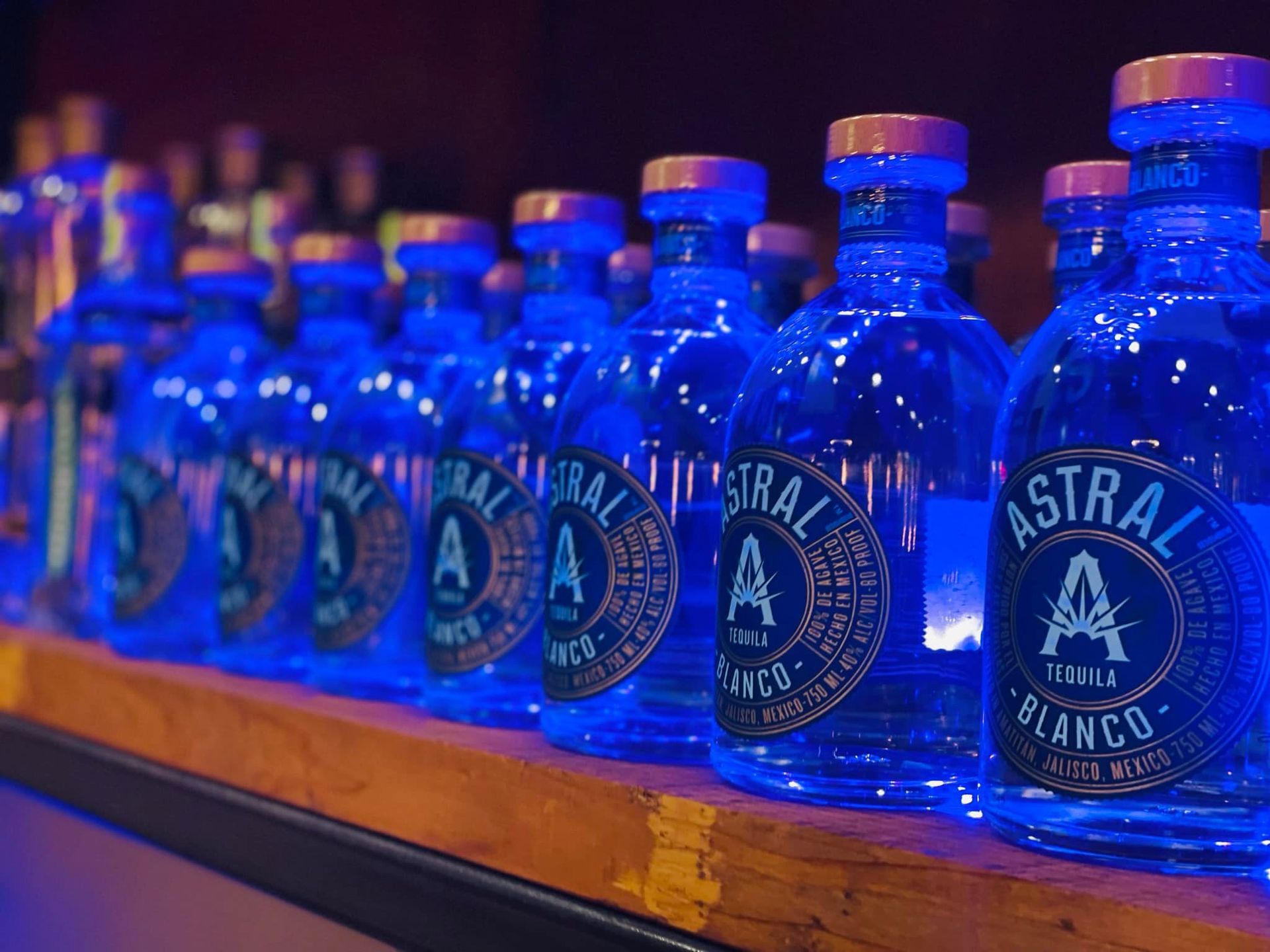 Row of blue Astral Blanco tequila bottles on a shelf, lit with purple and warm reflections