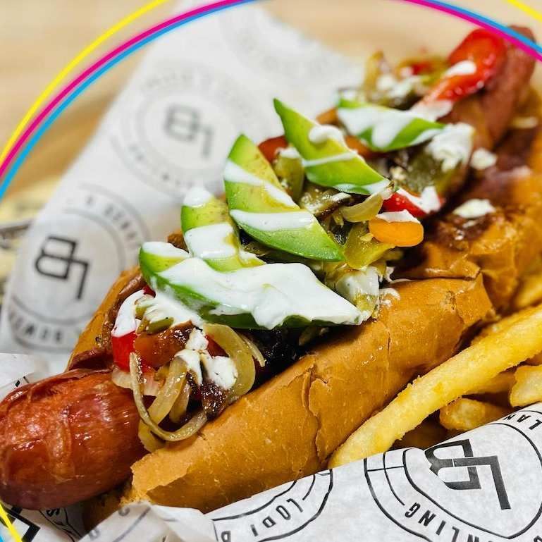 Loaded hot dog with avocado, onions, sauces, and fries on branded paper