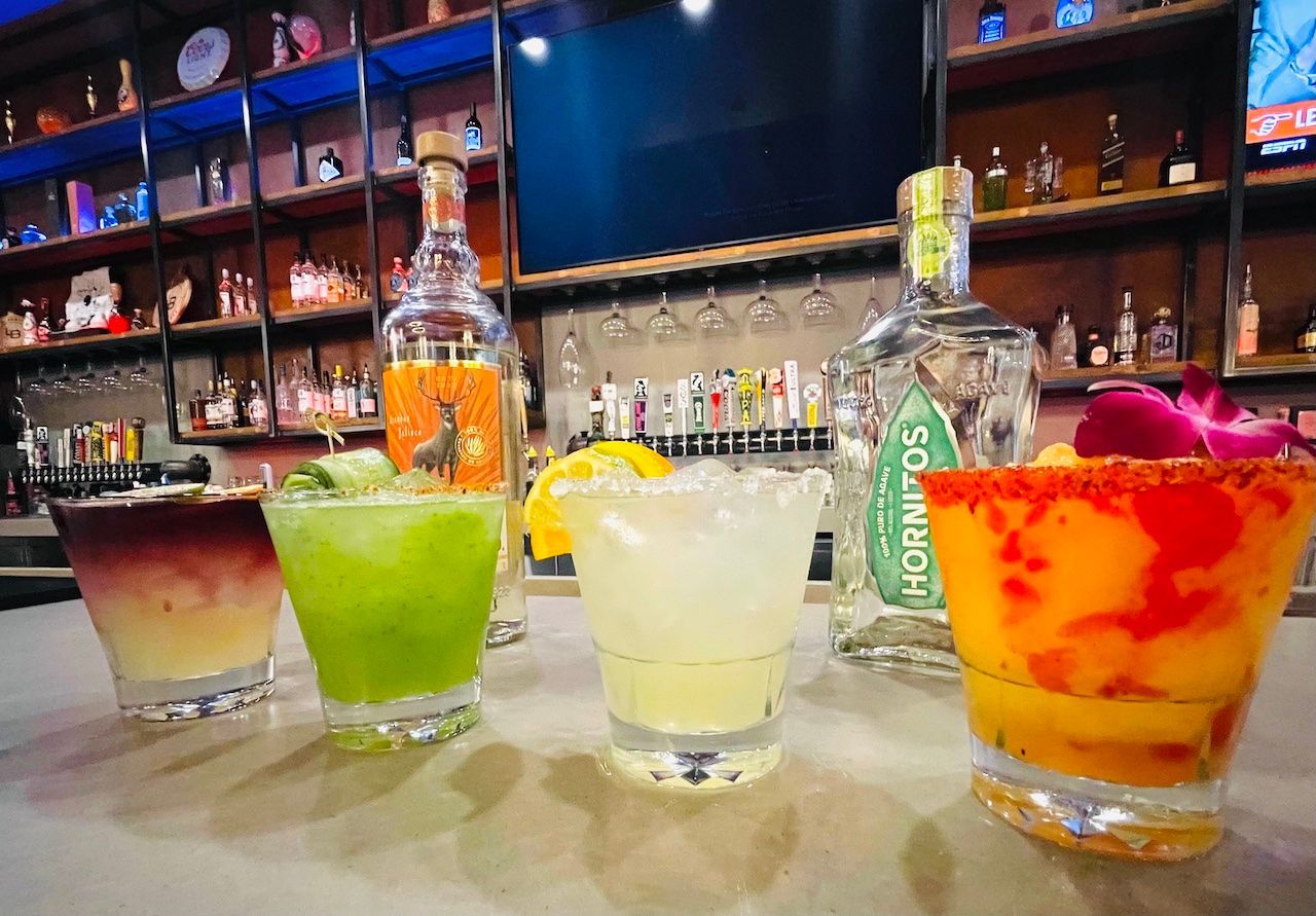 Four colorful cocktails on a bar counter, including green, yellow, and orange drinks with fruit garnishes.