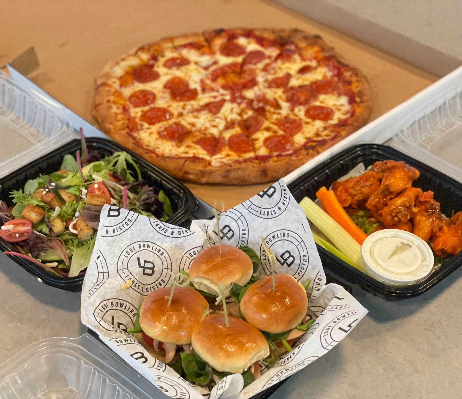 Pizza, sliders, salad, and buffalo wings with veggies and dip arranged in takeout trays on a table
