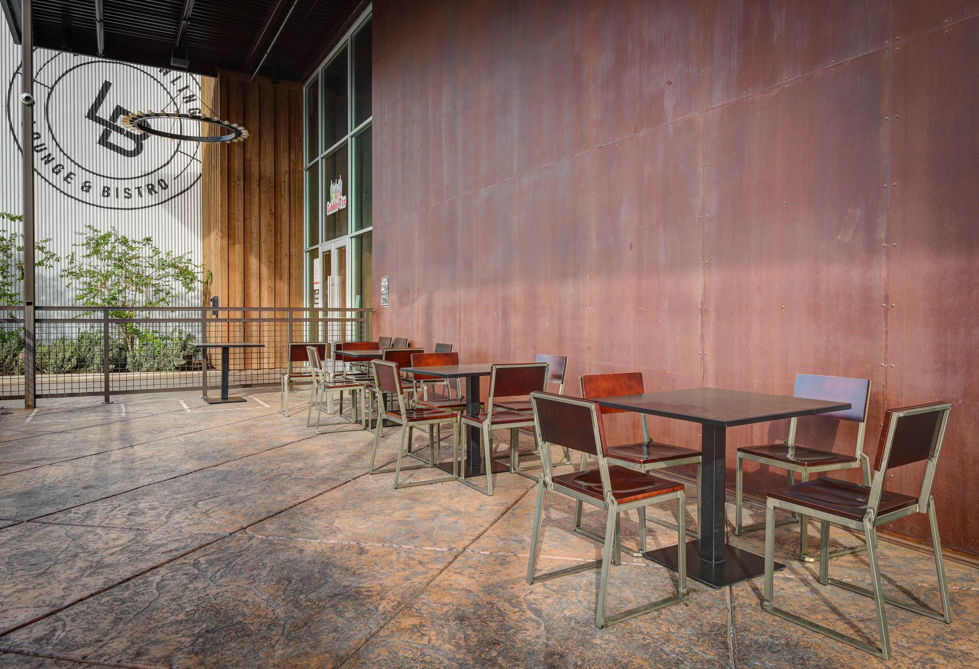 Outdoor café seating beside a large rust-colored wall with metal tables and chairs on a patterned floor
