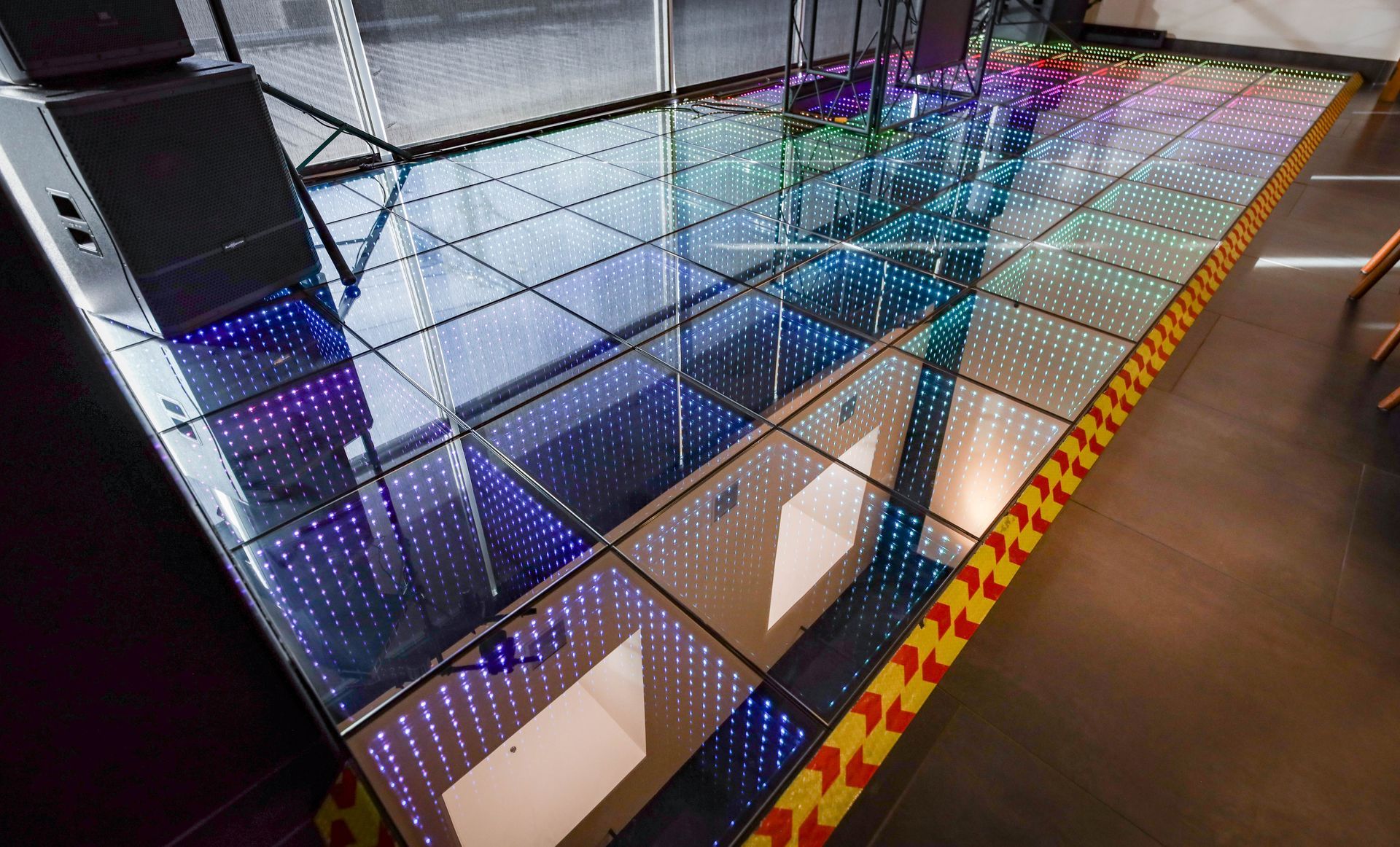 LED-lit glass floor walkway with a geometric grid pattern and orange-and-black caution edging in an indoor space