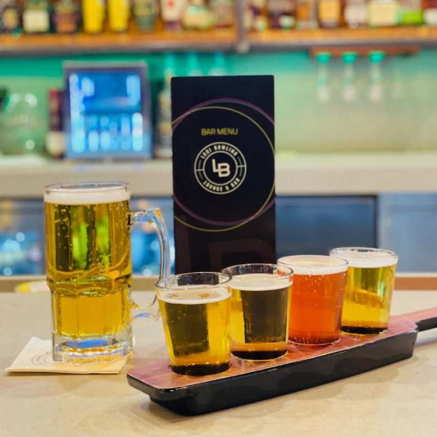 Flight of assorted beer samples on a bar counter with a large mug and bar menu sign