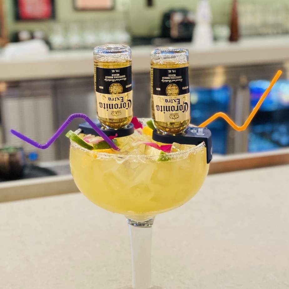 Yellow cocktail in a coupe glass topped with two miniature liquor bottles, with colorful stir sticks at a bar.