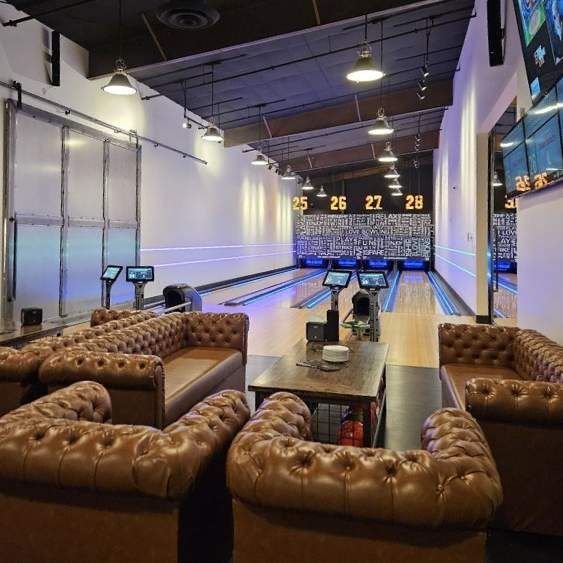 Bowling alley lounge with tufted leather couches facing illuminated lanes and scoring screens.