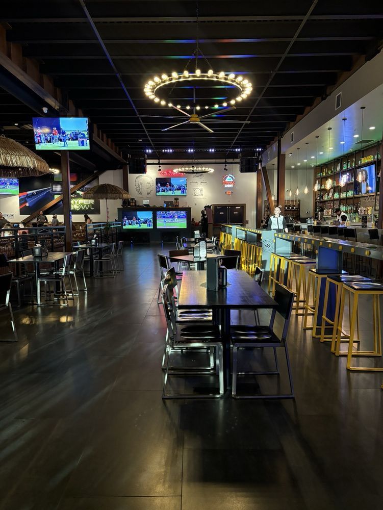 Dimly lit sports bar interior with TVs, a glowing ring chandelier, tables, and a long counter with stools.