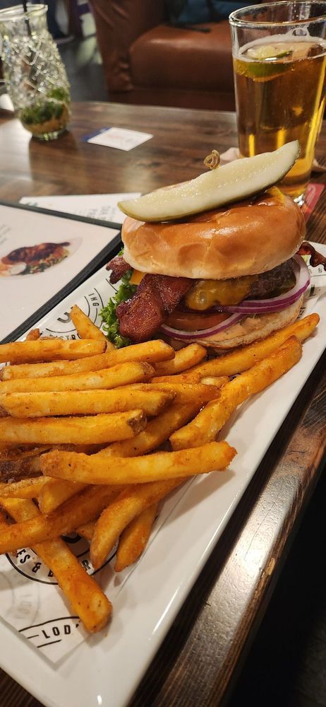 Burger and fries on a tray with a beer in a restaurant, with a pickle on top of the burger.