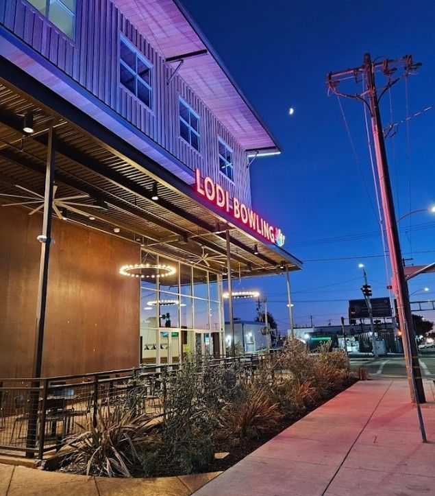Modern restaurant patio at dusk with neon sign, string lights, and sidewalk beside a utility pole