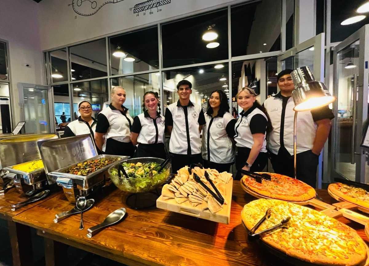 Caterers standing behind a buffet table with salads, sandwiches, and pizzas in a bright indoor venue