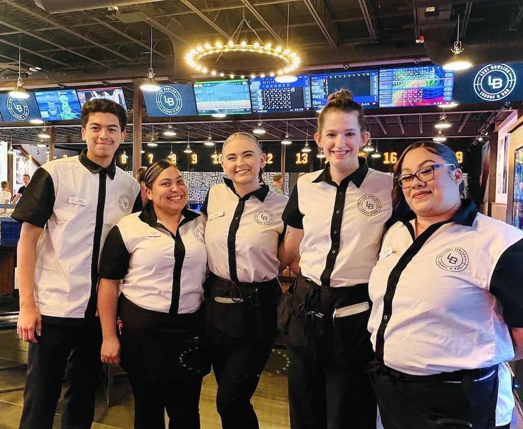 Five smiling staff members in matching white shirts pose inside a brightly lit arcade or bar.