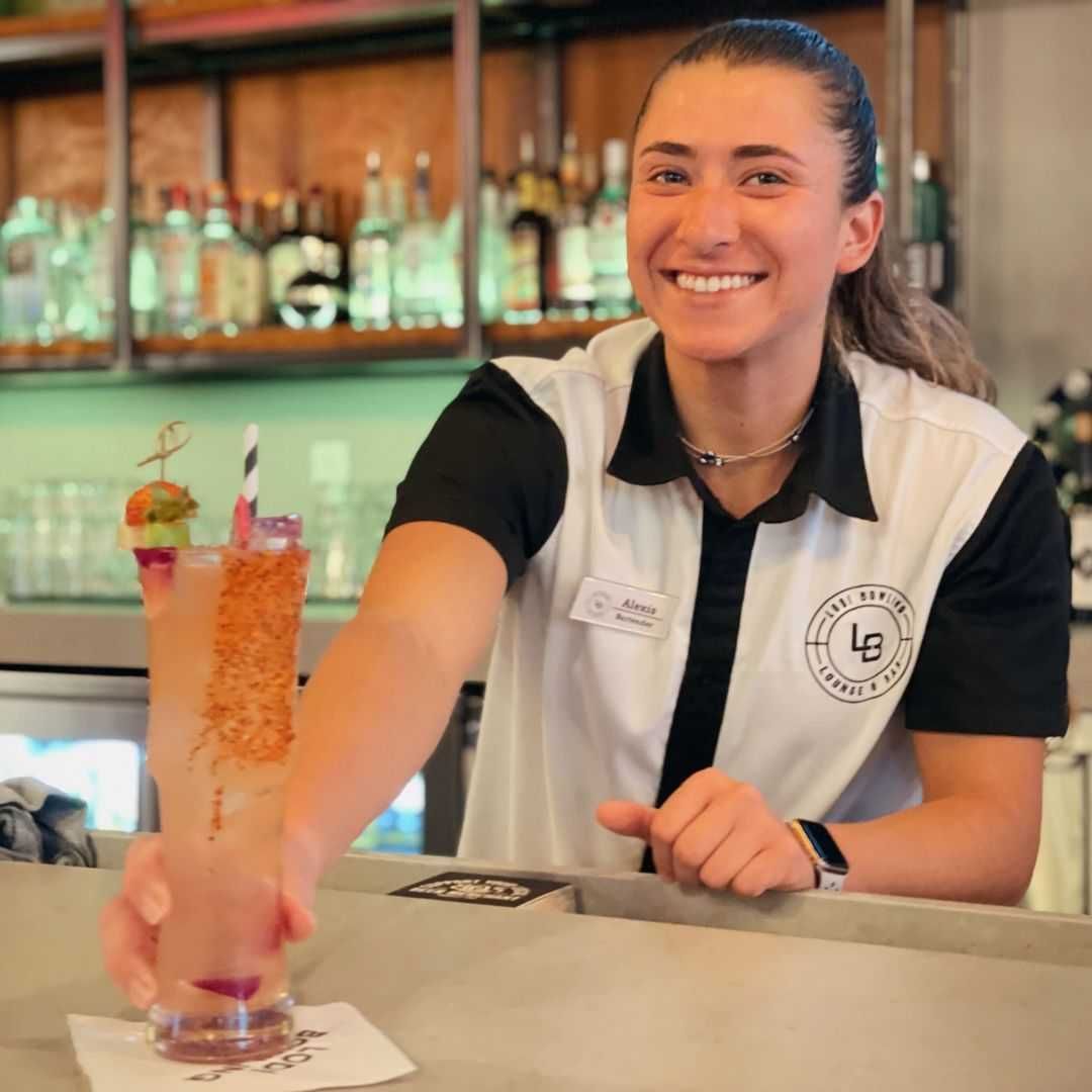 Smiling bartender handing a tall pink cocktail across a bar counter
