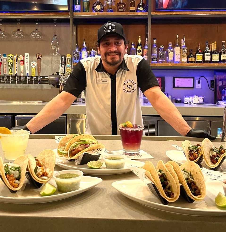 Bartender smiling behind a counter with tacos and a red drink in a lively bar