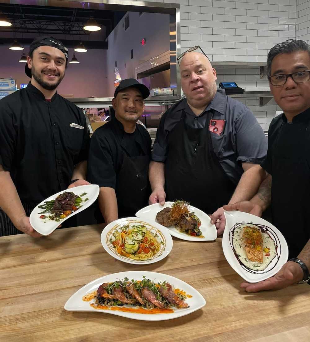 Four restaurant staff posing with plated dishes around a wooden table in a kitchen area