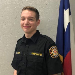 A young man in a firefighter uniform is standing in front of a flag.