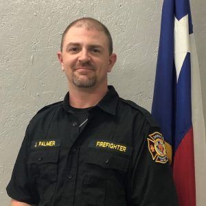 A man in a firefighter uniform is standing in front of a flag.