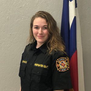 A woman in a firefighter uniform is standing in front of a flag.