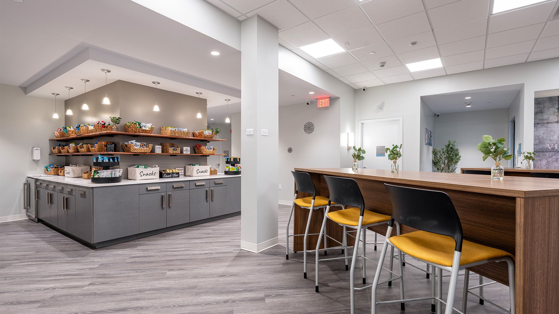 A bright office breakroom featuring a snack bar, grey cabinets, and a wooden counter with yellow-seated stools.