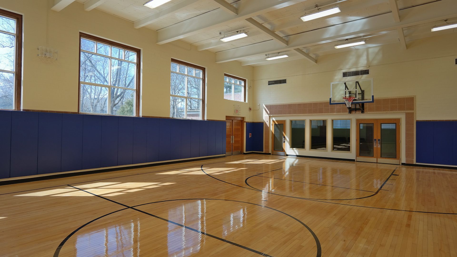 Empty indoor basketball court with shiny hardwood floors, blue wall padding, large windows, and a basketball hoop.
