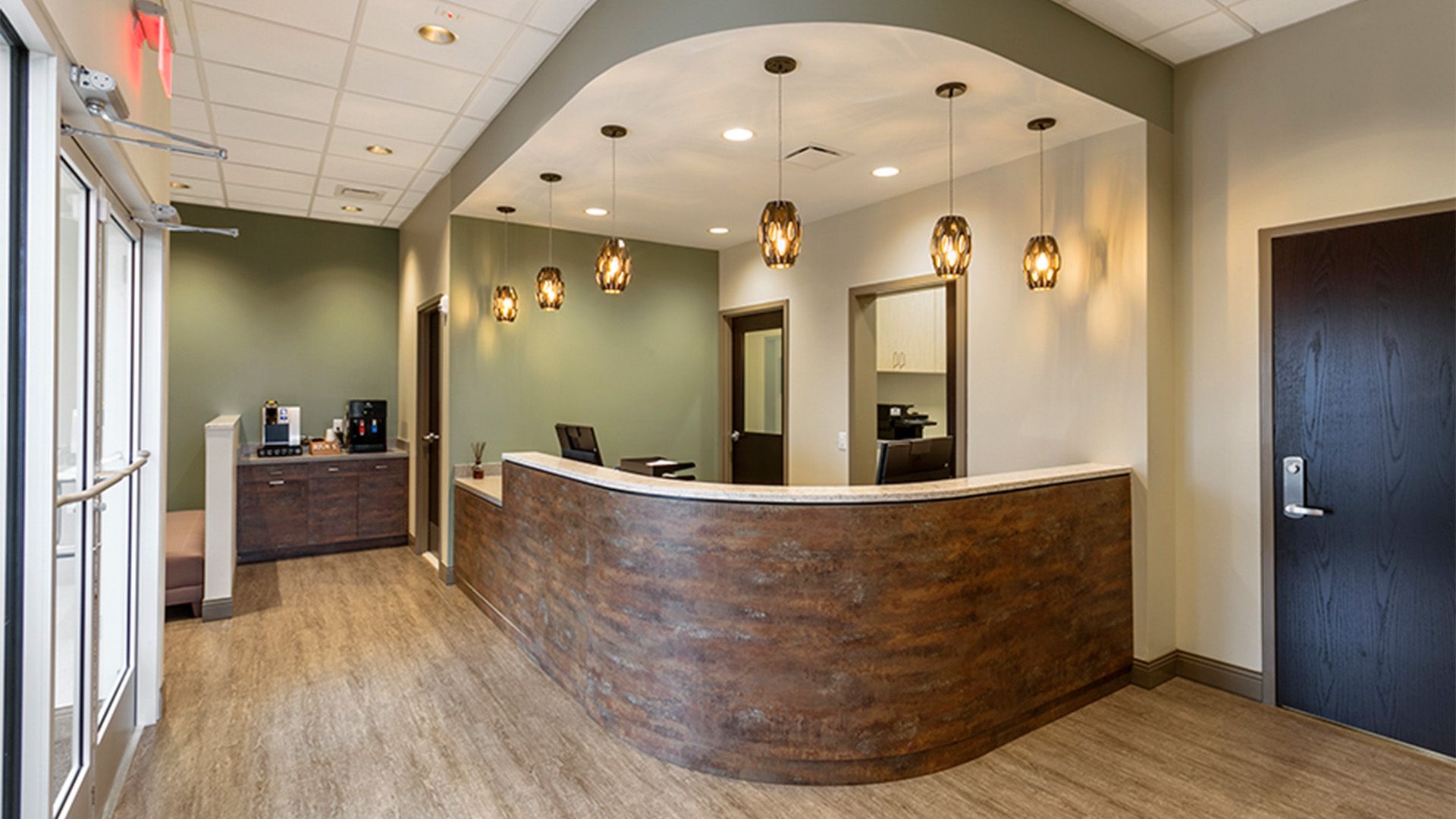 A brightly lit reception area with a curved wood-panel desk, pendant lights, and sage green accent walls.