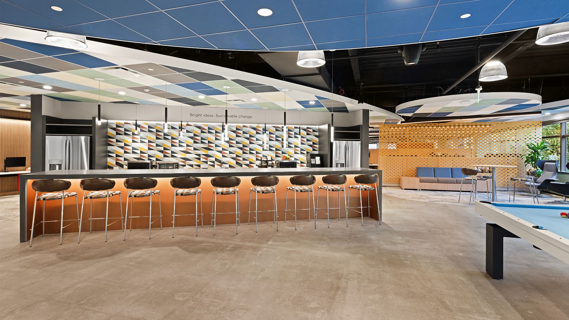 Modern office lounge with a long bar, wooden stools, decorative wall tiles, a pool table, and blue patterned ceiling panels.