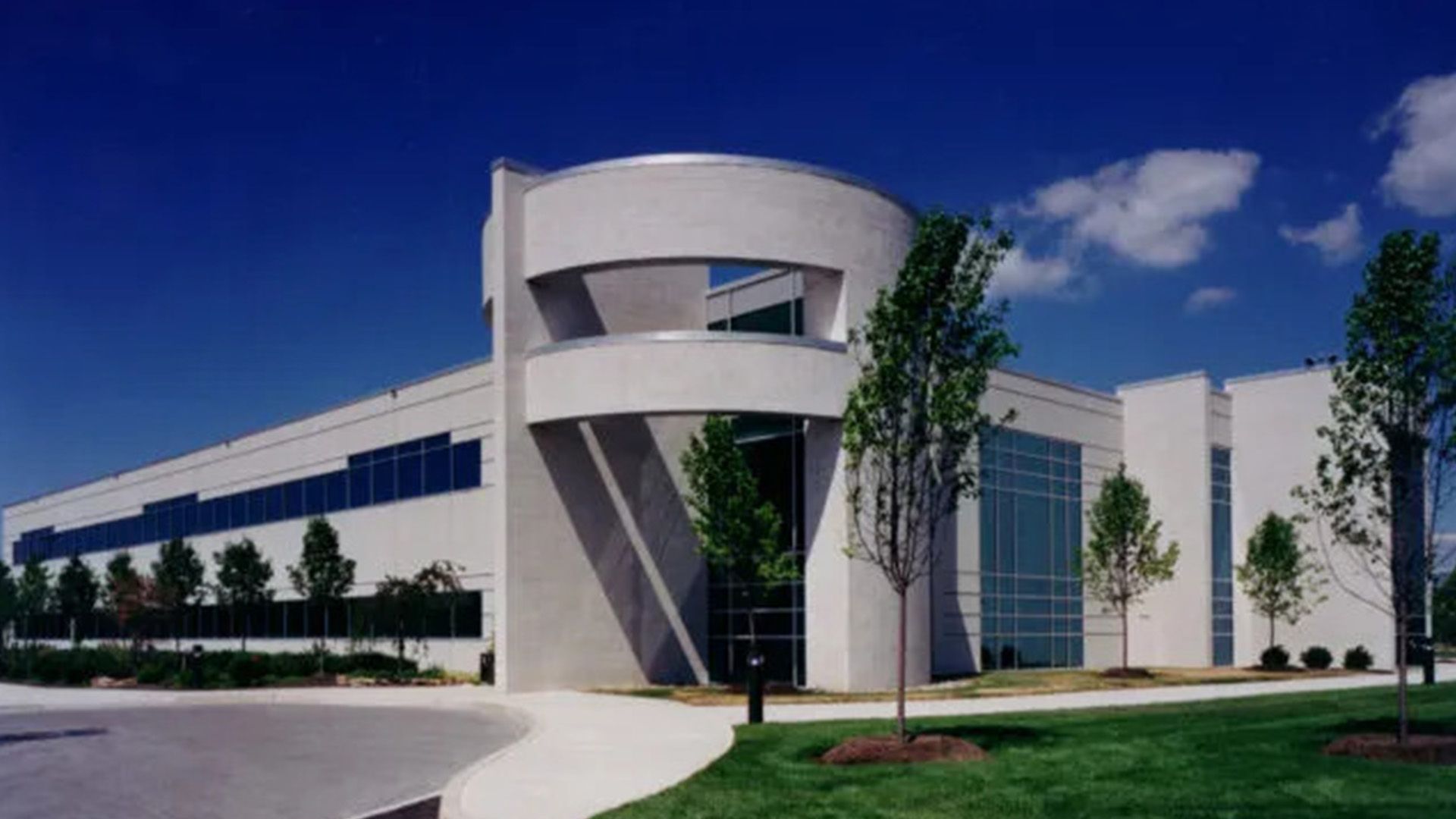 A modern white office building with a curved glass entrance tower, surrounded by a lawn and young trees under a blue sky.