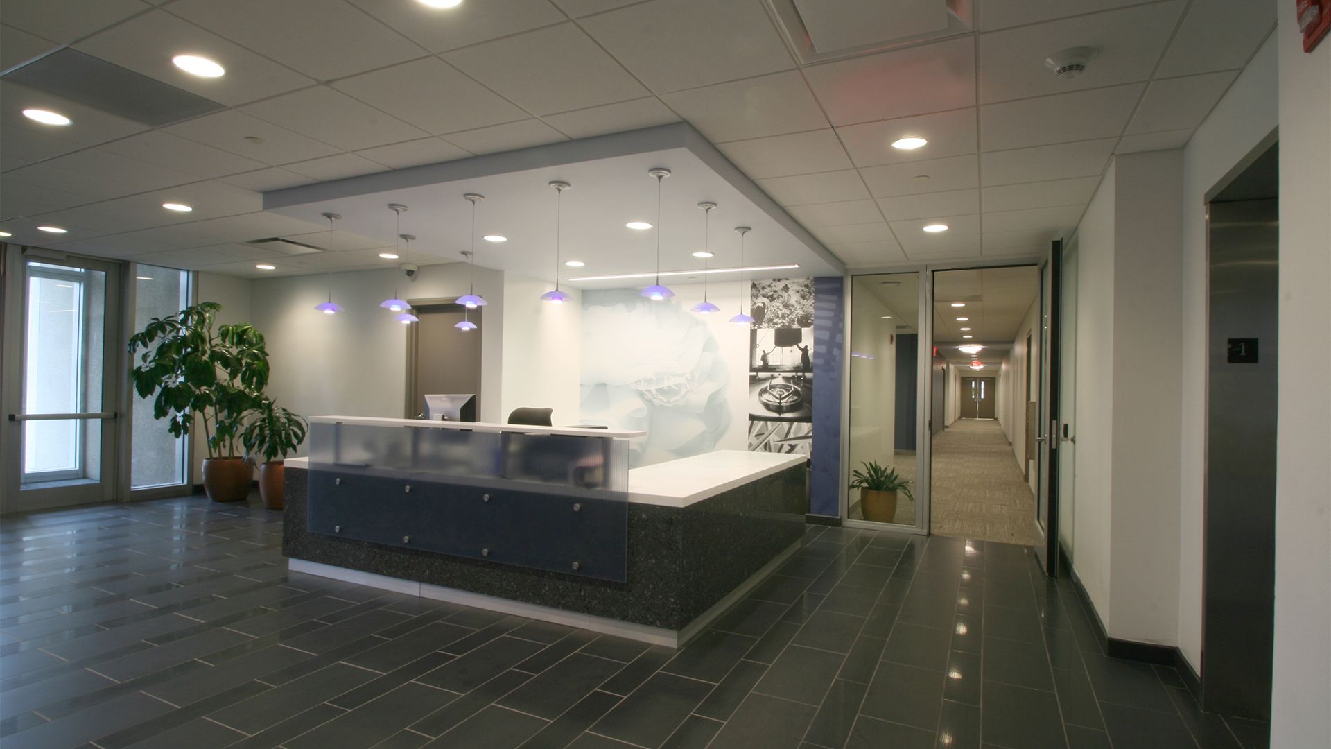 A modern office lobby with a reception desk, gray tile floors, pendant lighting, and a view into a long hallway.
