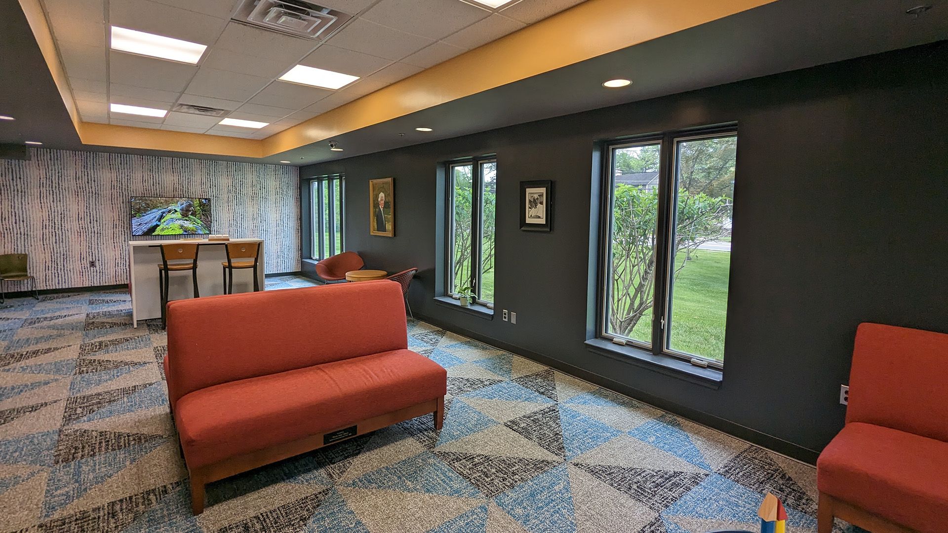 A modern lounge area with patterned blue carpet, orange seating, and floor-to-ceiling windows looking out to greenery.