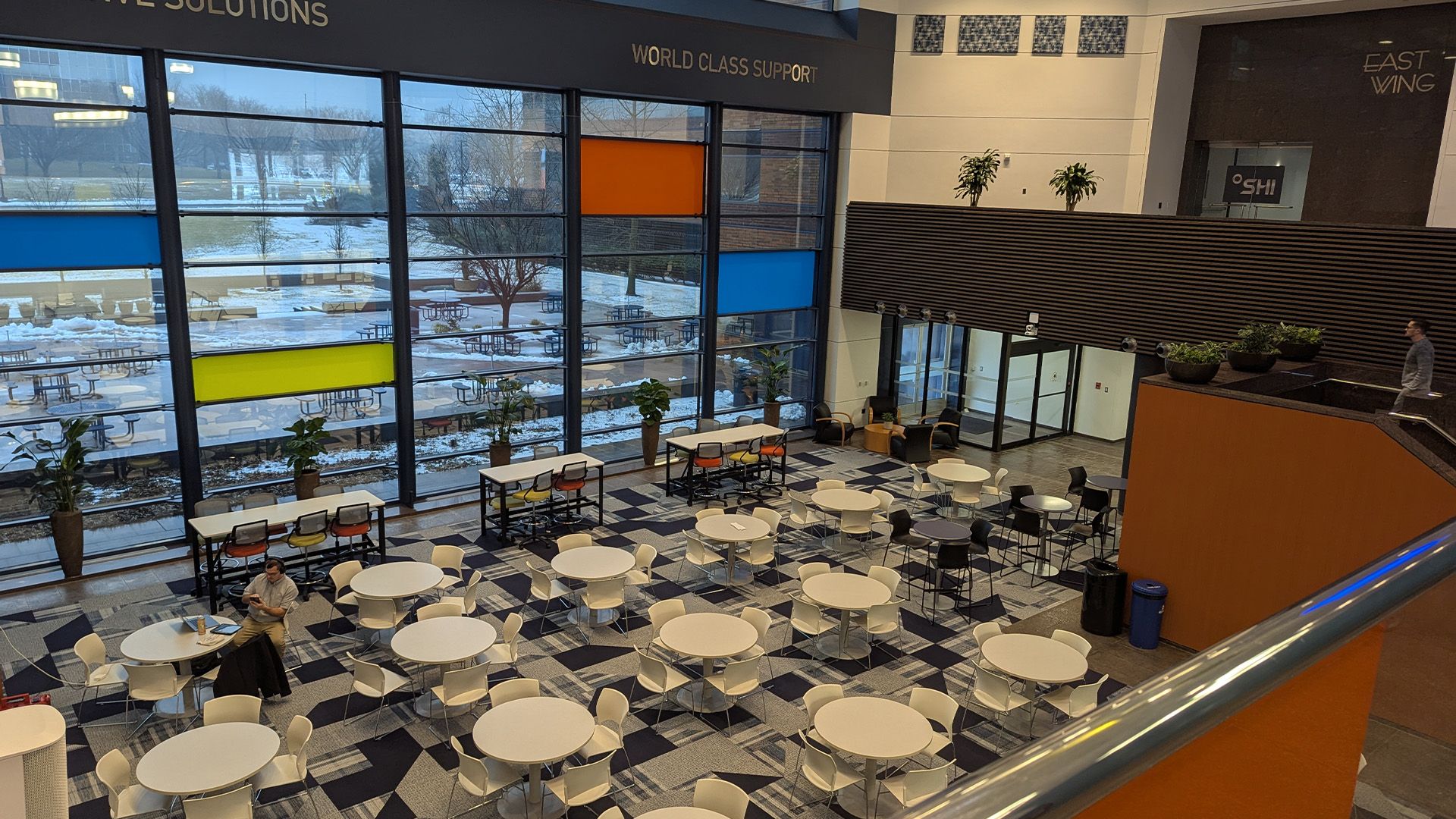 A high-angle view of a modern, multi-story cafeteria with circular white tables and chairs, large windows, and a snowy view.