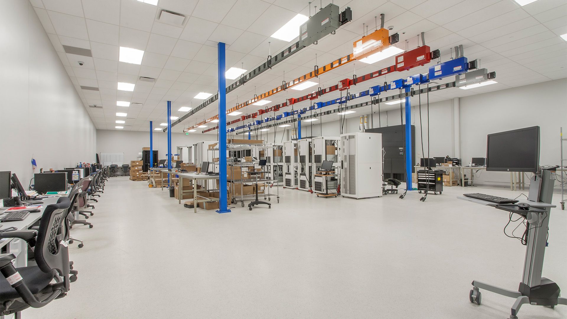 A bright, modern laboratory or industrial workspace with rows of equipment, work tables, and overhead utility lines.