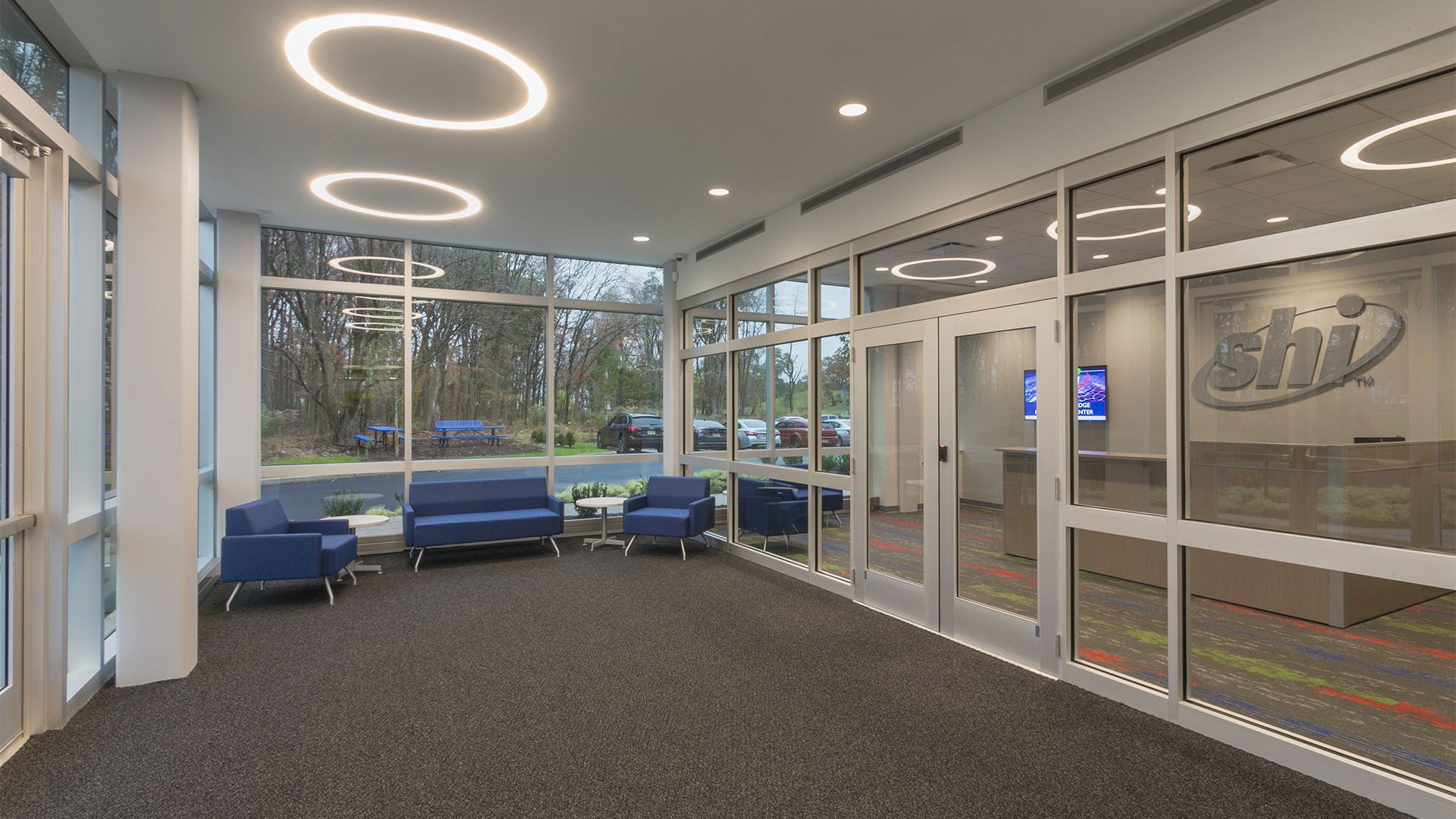 Modern office reception area with blue chairs, floor-to-ceiling windows, and a SHI company logo on a glass wall.