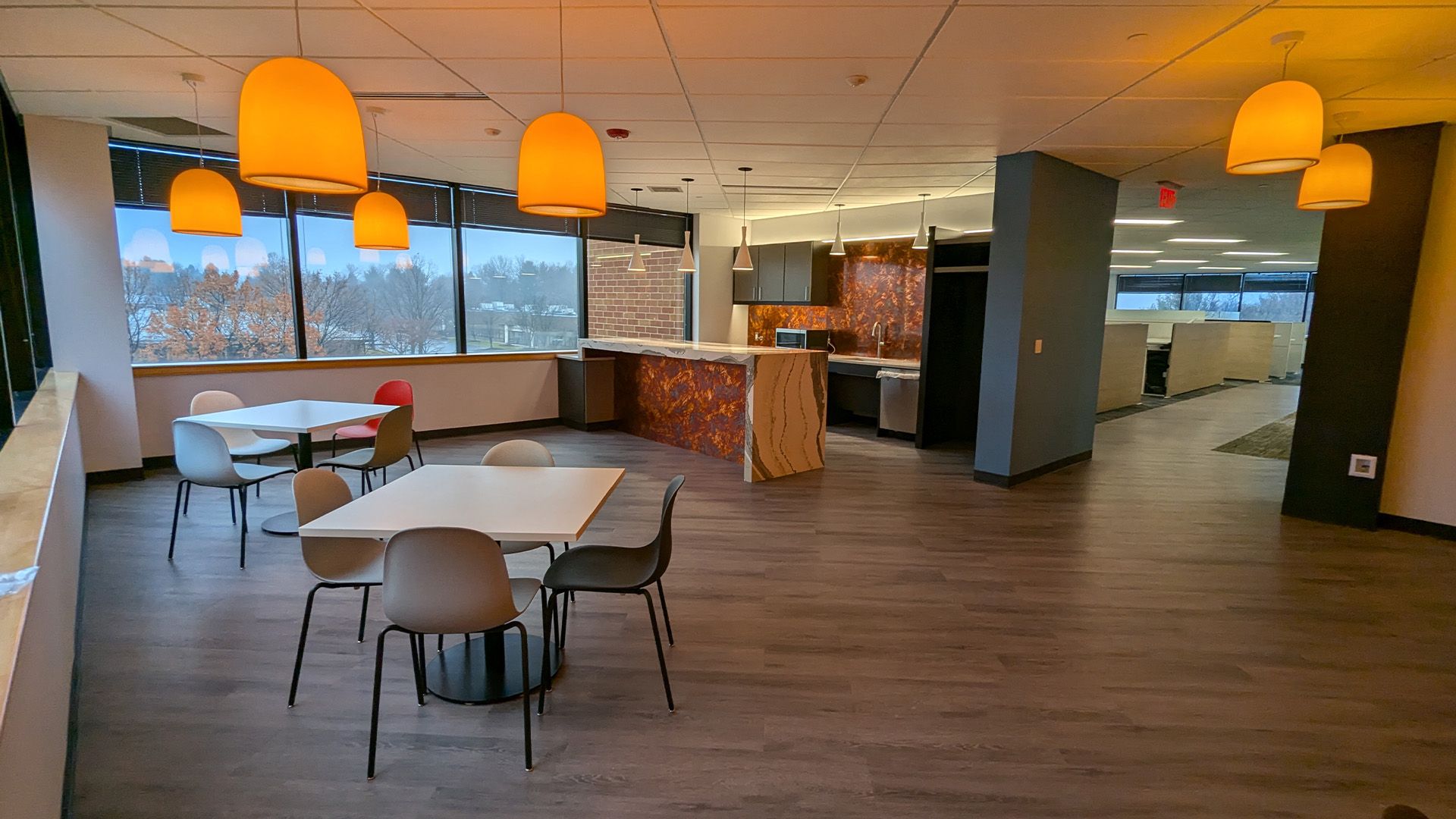 A modern office break room with two tables, chairs, pendant lights, and a small kitchen area with a wood-patterned counter.
