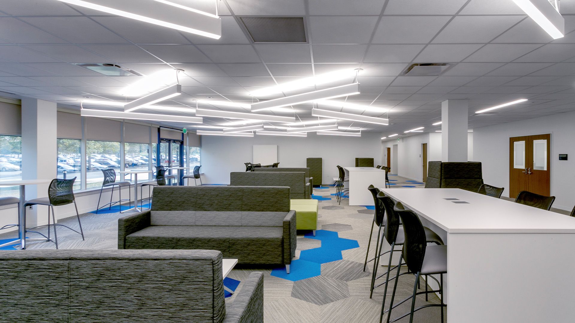 Modern office lounge with gray couches, a tall white workspace, chairs, and unique geometric overhead lighting.
