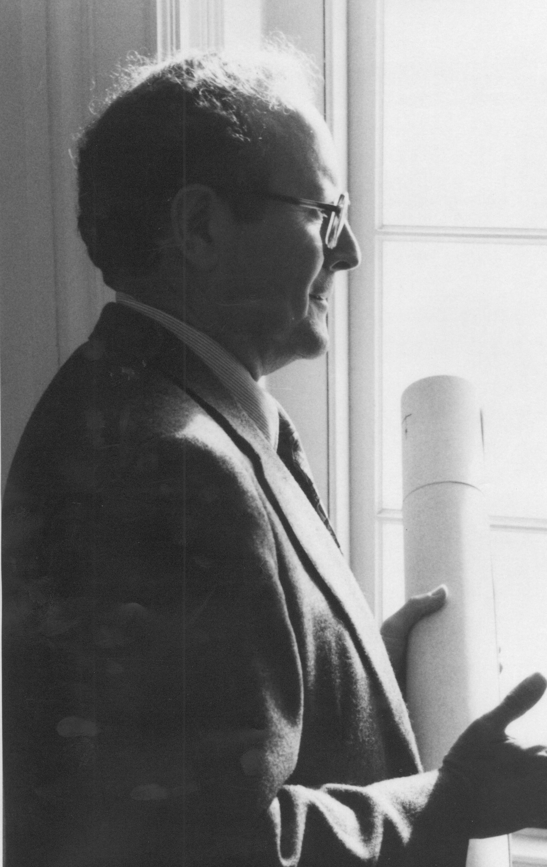 A person in a suit stands by a window, holding a rolled document and gesturing while speaking.