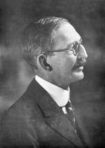 A profile portrait of a man in a dark suit, tie, and glasses, looking to the right against a dark, blurred background.