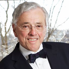 A person in a suit and bow tie smiles warmly against a blurred, bright background of outdoor trees.