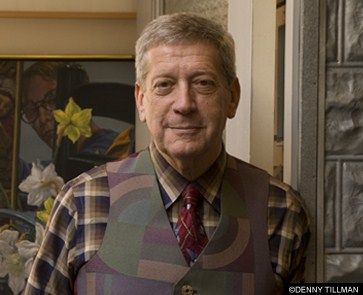 A person in a plaid shirt and patterned vest, posed indoors with floral artwork in the background.