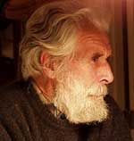 A side profile portrait of a person with wavy gray hair and a long, thick white beard, wearing a dark sweater.